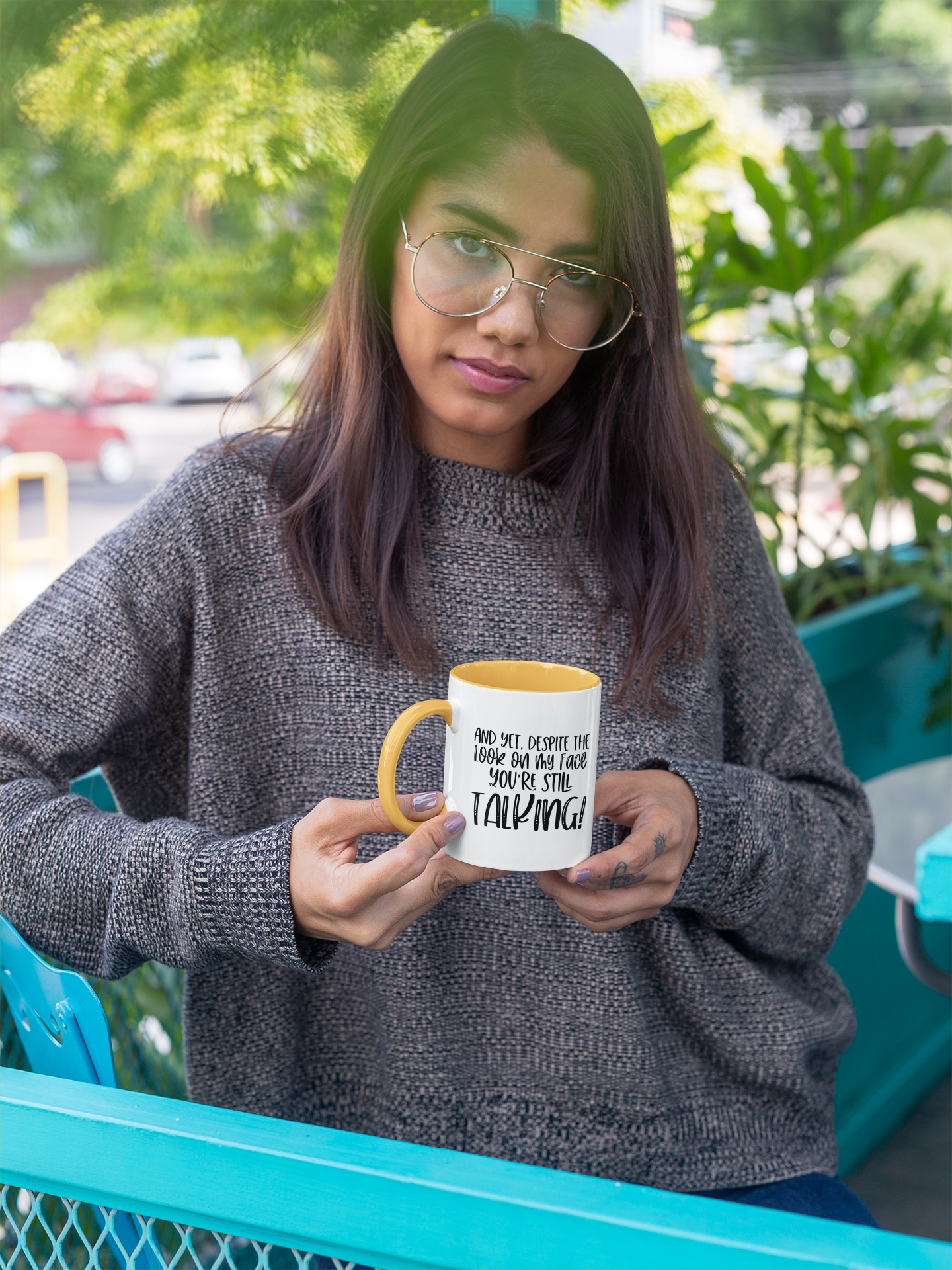 A person holding a white ceramic mug with text on it, sitting outdoors with plants in the background.