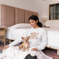 Woman sitting on a bed with a small dog, wearing a sweater with colorful text that says Hey Sugar Pie, in a bedroom setting.