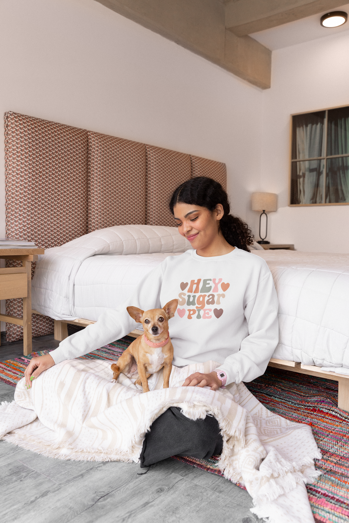 Woman sitting on a bed with a small dog, wearing a sweater with colorful text that says Hey Sugar Pie, in a bedroom setting.