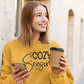 Woman in a yellow sweatshirt with 'Cozy Season' text, holding a phone and a coffee cup outdoors.