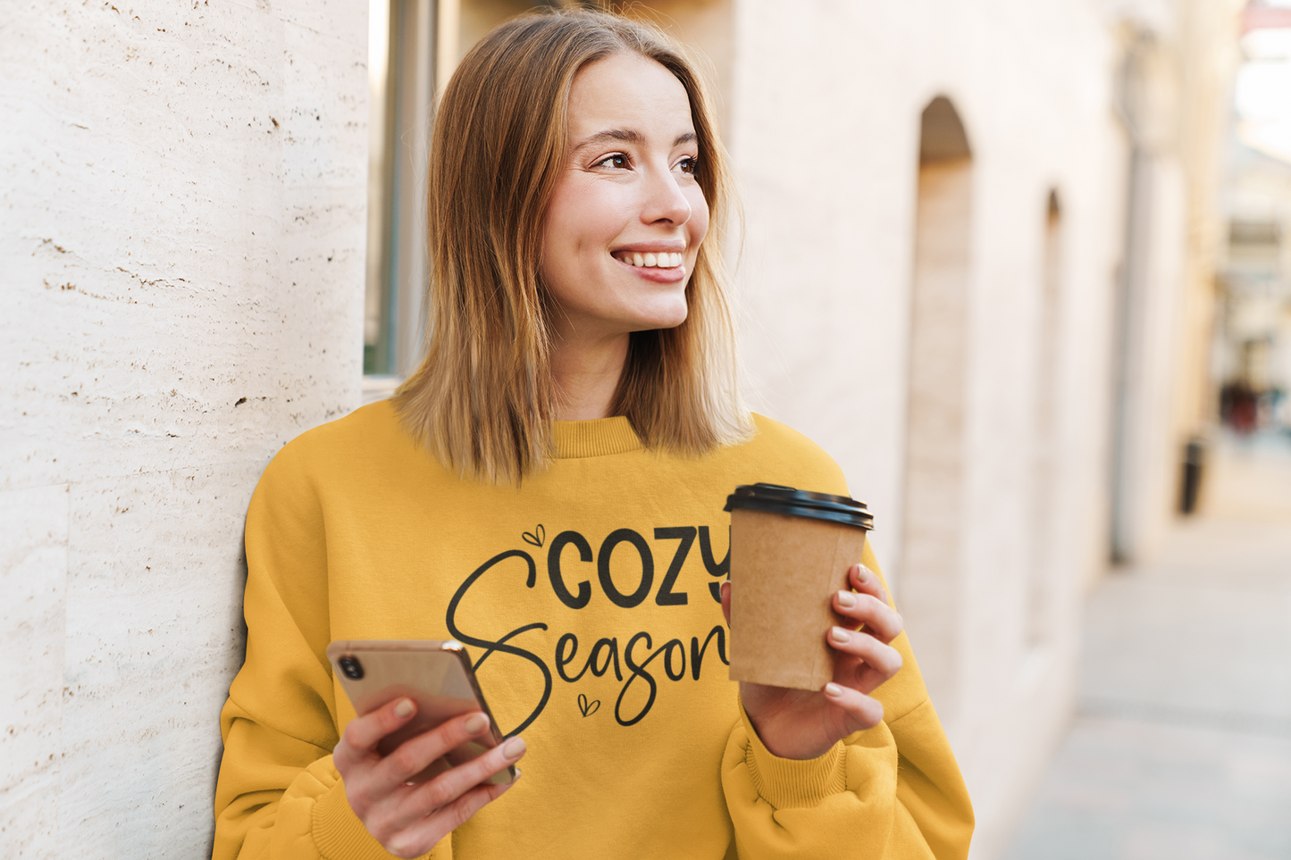 Woman in a yellow sweatshirt with 'Cozy Season' text, holding a phone and a coffee cup outdoors.