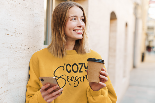 Woman in a yellow sweatshirt with 'Cozy Season' text, holding a phone and a coffee cup outdoors.