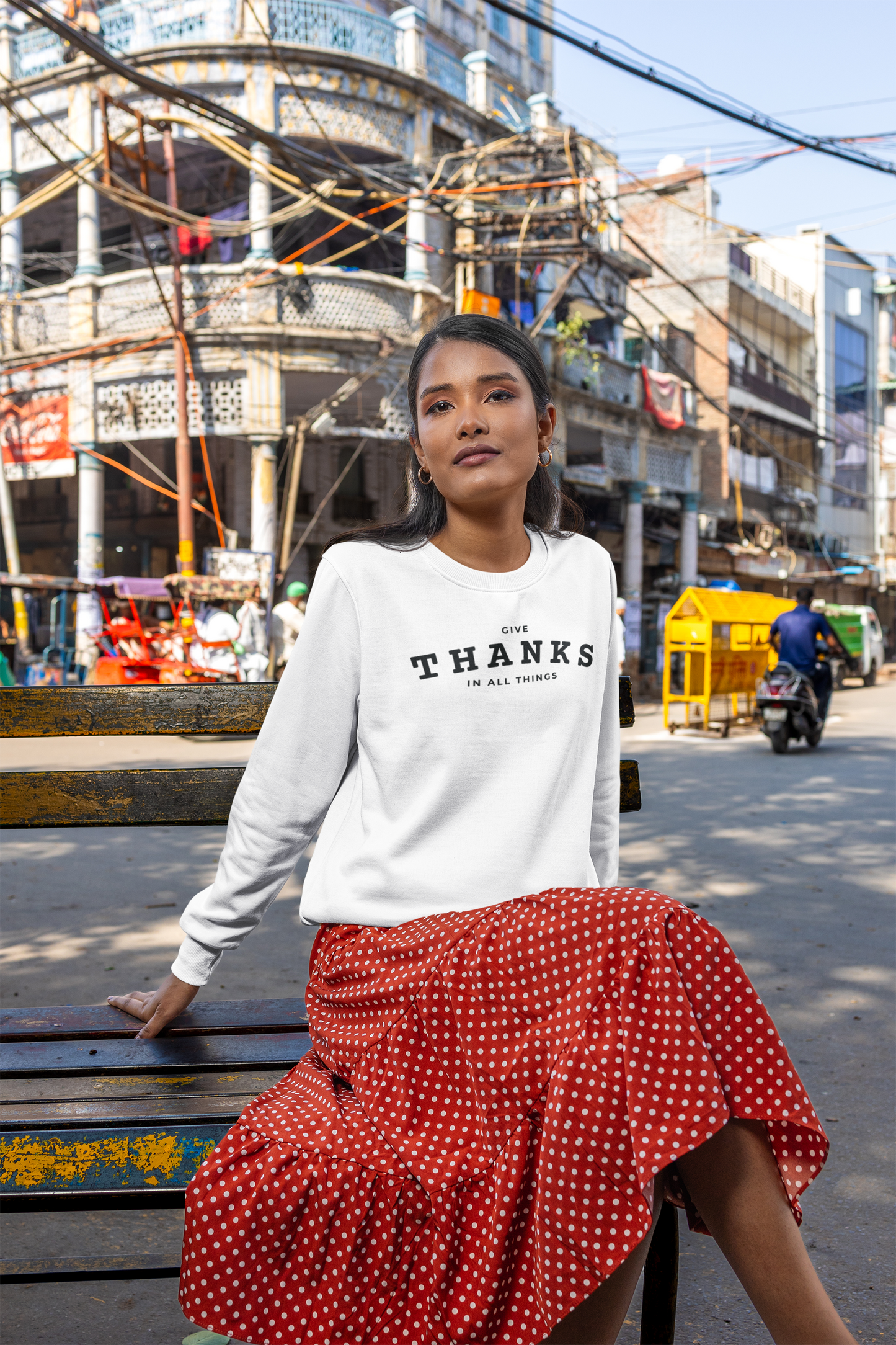 Woman sitting on a bench wearing a white sweatshirt with text and a red polka dot skirt in an urban setting.